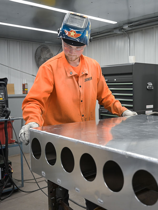 metal-fabricator PAVE Manufacturing team member in orange coat and welders helmet working on a metal project