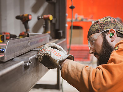 An fabricator in safety glasses and hearing protection carefully measuring or marking steel unistrut on a workbench in a shop.